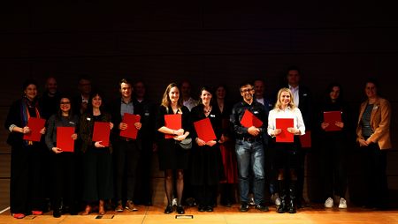 Eine Gruppe von Menschen steht auf einer B&uuml;hne und h&auml;lt rote Ordner in den H&auml;nden, l&auml;chelnd und stolz.